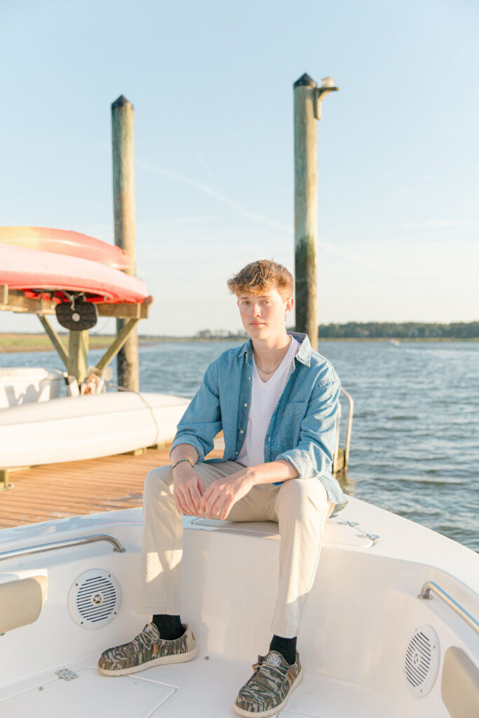 A young man in a chambray shirt sitting on the bow of his boat on the May River next to a dock at golden hour in Bluffton, SC for his senior portrait session with Bluffton senior photographer