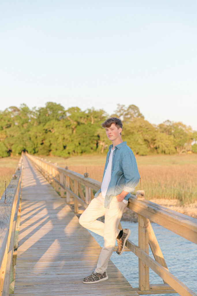 A young man in a chambray shirt standing on a long dock on the May River in Bluffton SC for his senior portrait session with Bluffton SC Photographer
