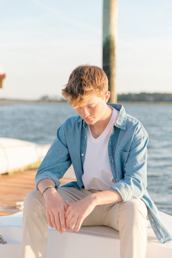 A young high school aged man sitting on his boat looking down at his hands next to the dock on the May River during his senior portrait session with Bluffton senior photographer