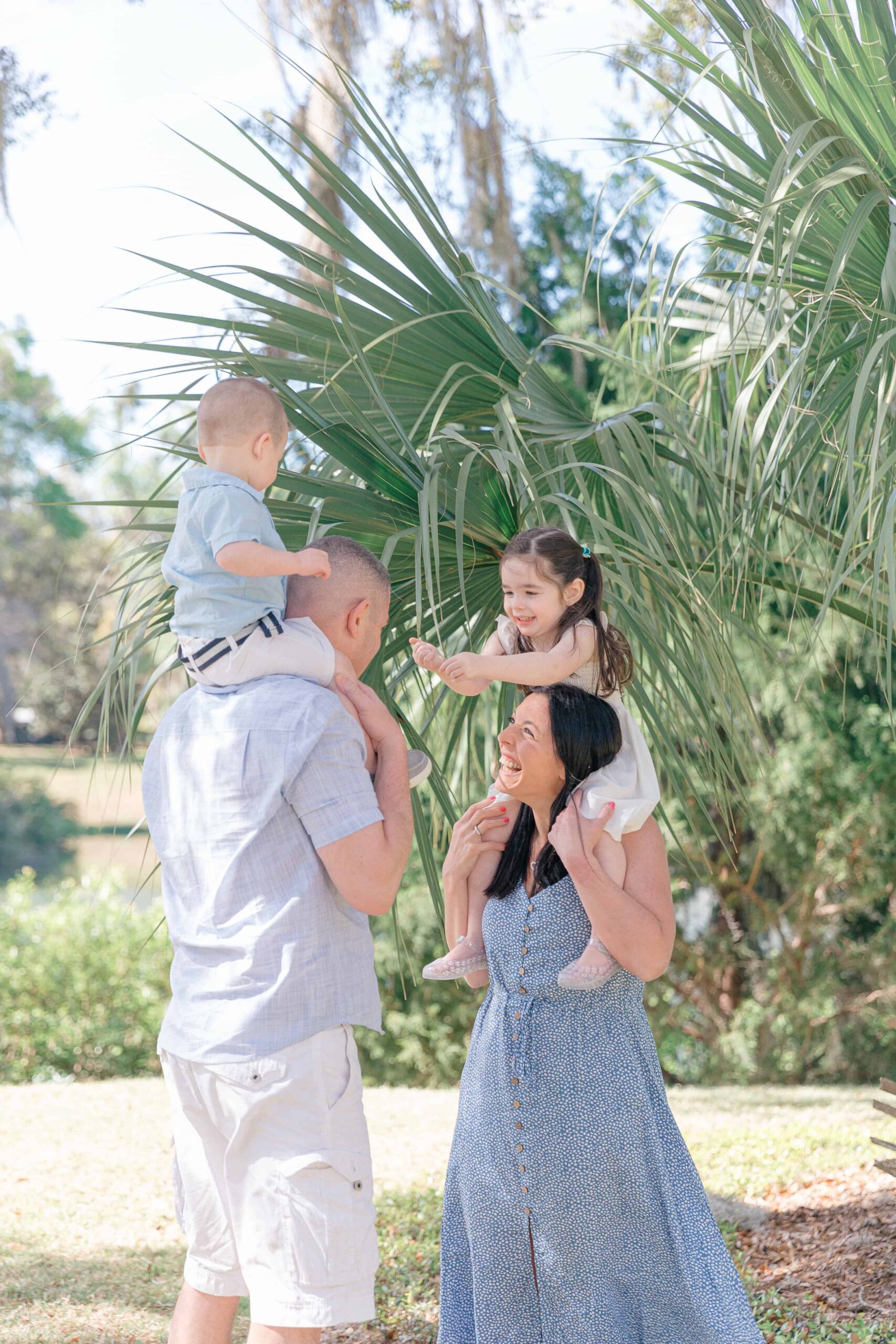 A man and a woman stand outdoors, each carrying a child on their shoulders. The woman looks up, smiling, while the man looks at the child on his shoulders. They are surrounded by greenery.