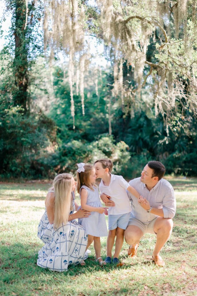 Little boy kissing his sister on her cheek under an oak tree with mom and dad admiring them with Hilton Head Family Photographer, Lamp & Light Photography.